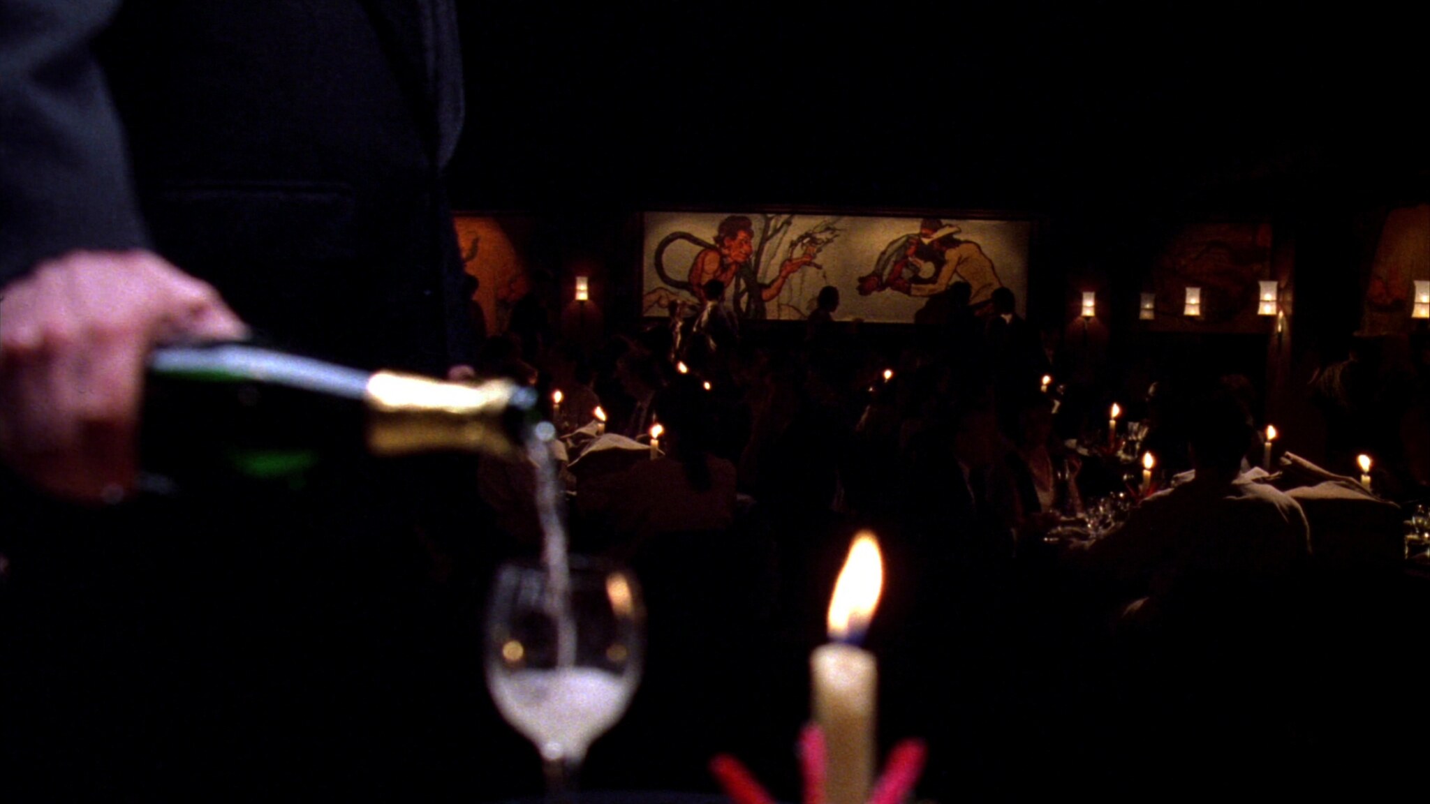 A candlelit dining room filled with guests as warm blurred silhouettes, champagne pouring in the foreground