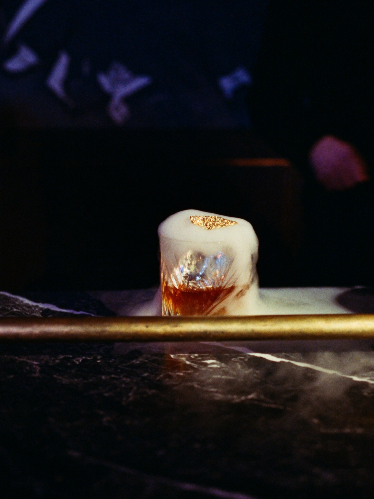 A crystal rocks glass of amber old fashioned with one clear cube on a dark marble bar
