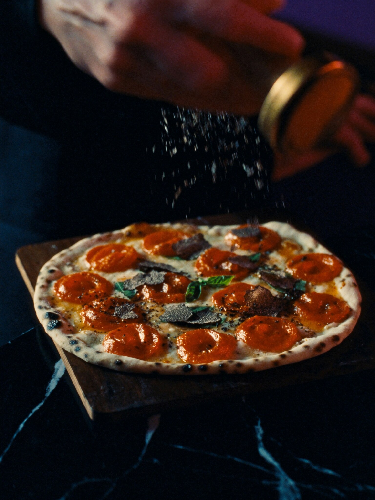 A leopard-spotted pizza with pepperoni cups and truffle shavings falling through the light