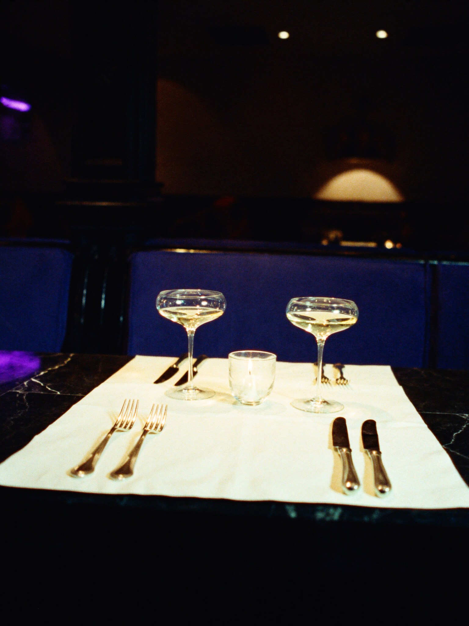 A two-top set at a navy velvet banquette with two champagne coupes and a single candle