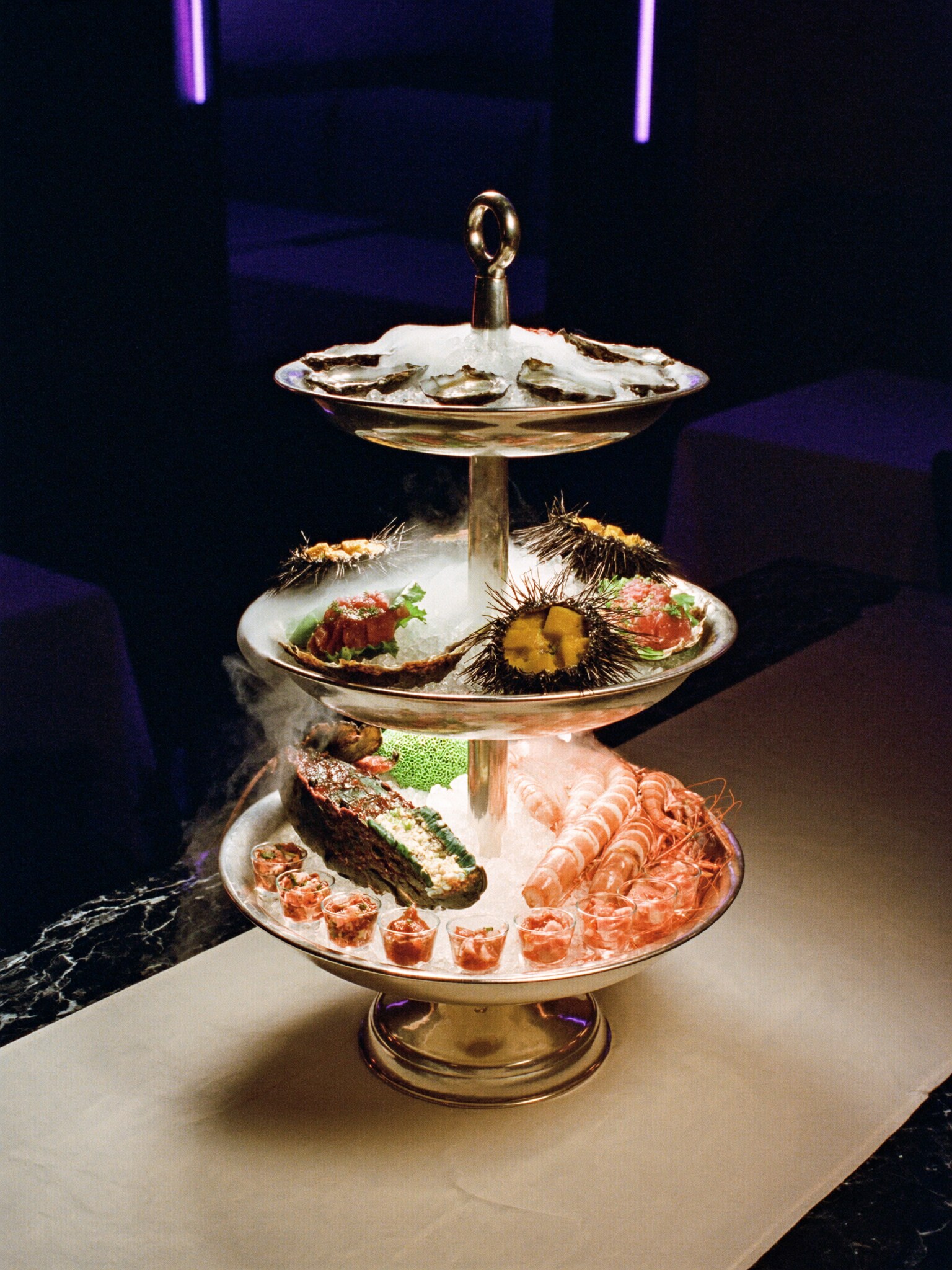 A three-tier silver seafood tower on crushed ice with dry-ice smoke rolling down the tiers